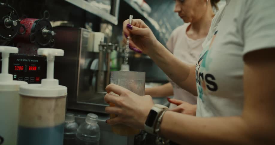 A barista stirs ingredients in a mixing cup while preparing a beverage in a busy cafe kitchen