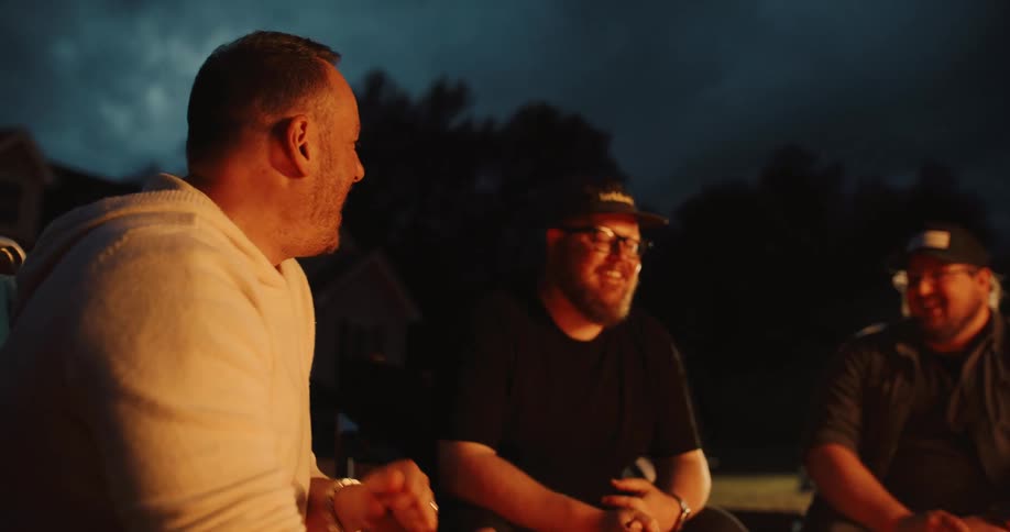 A group of male friends share stories and laughter while gathered around a cozy campfire at dusk