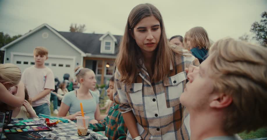 A young woman smiles while carefully applying face paint to a young man at an outdoor community event