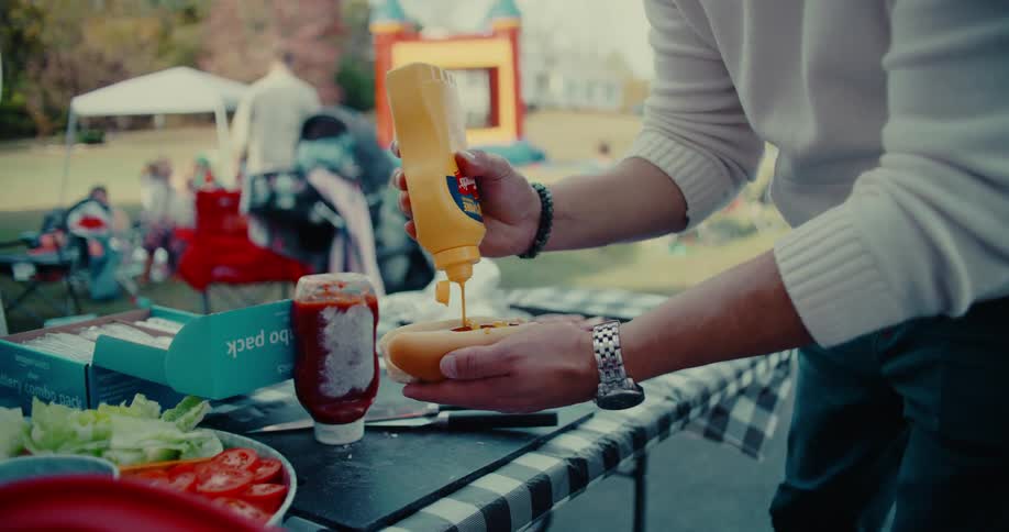 A man prepares a hot dog with mustard in slow motion at a casual outdoor party before taking a bite