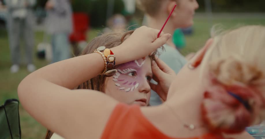 A makeup artist carefully paints a beautiful purple and white design on a young girl's face at an outdoor party