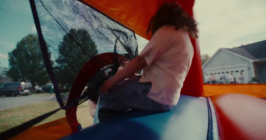 A happy mother watches her daughter playfully jump up and down inside a colorful bouncy house during a sunny day in slow motion