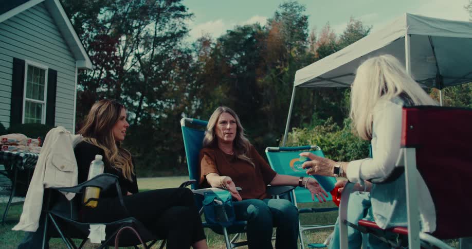 Three women have a casual conversation while sitting in lawn chairs at a sunny backyard get-together
