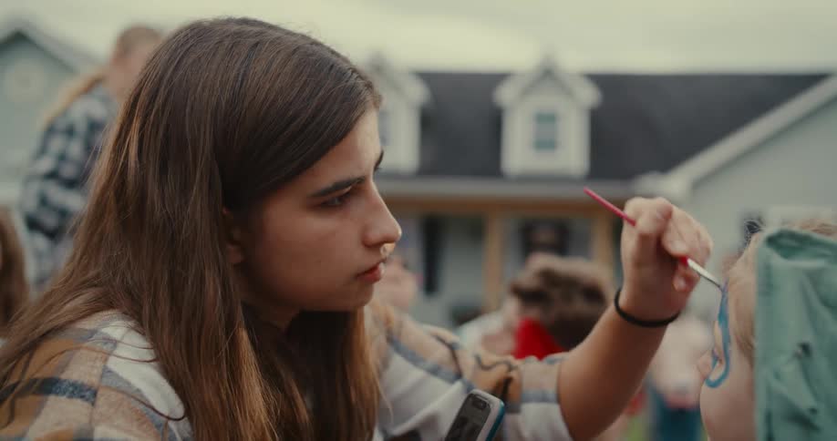 A focused young woman carefully applies face paint to a child's cheek at an outdoor party