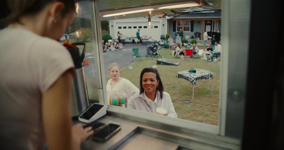 A happy woman receives her drink from a food truck and pays with a card during a fun neighborhood block party