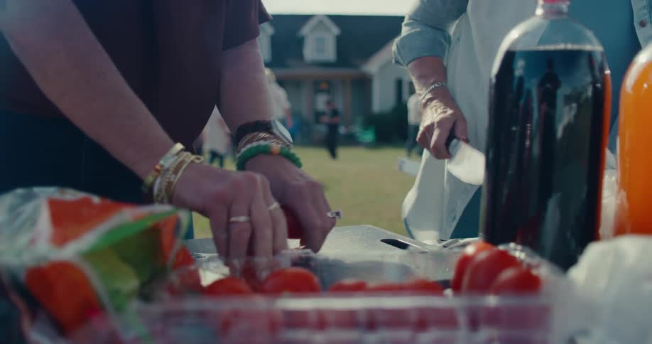 Two women slice fresh red tomatoes on a cutting board while preparing food for an outdoor barbecue party in a suburban backyard