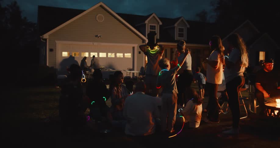 A diverse group of adults and children enjoy a fun nighttime party in a suburban front yard, adorned with colorful glow sticks