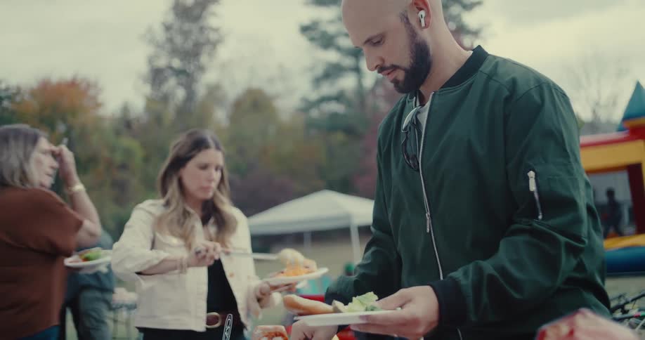 A bearded man in a green jacket carefully prepares his plate at an outdoor community barbecue in slow motion