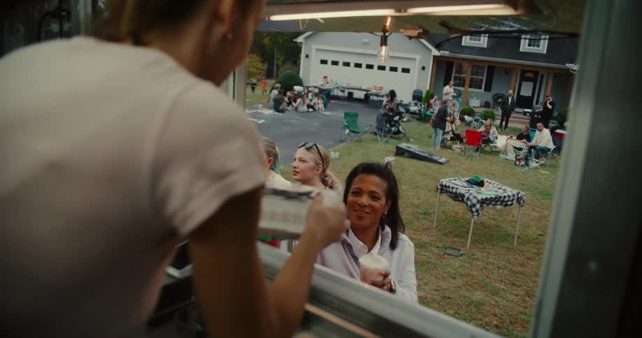 A smiling woman receives her food and drink order from a food truck employee during a lively neighborhood block party in a suburban setting