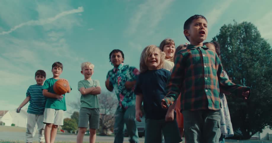 A diverse group of excited children cheer and then run together in slow motion on a sunny day