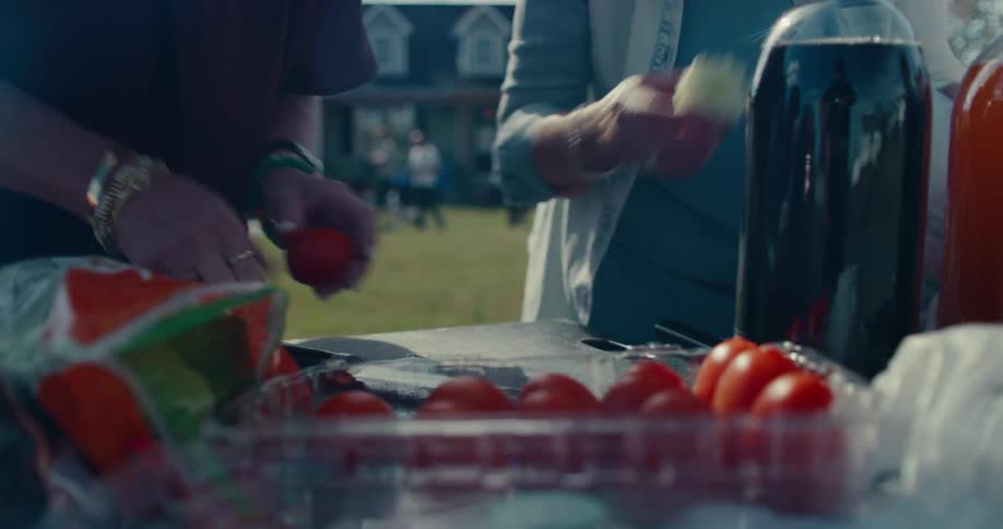 Two women slice fresh tomatoes together on a cutting board while preparing food for a sunny outdoor backyard gathering
