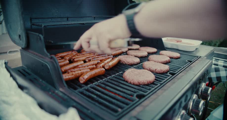 A person slowly lifts the lid of a grill and uses tongs to turn hot dogs cooking next to hamburgers at a backyard barbecue