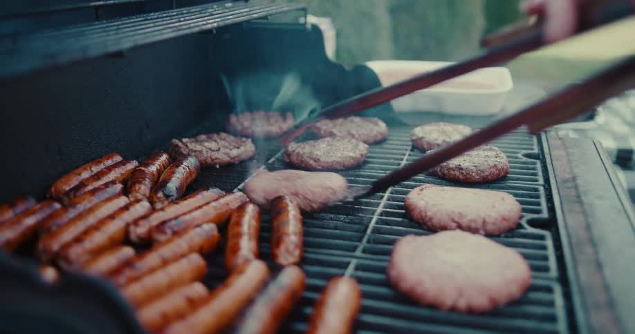 A close-up slow-motion shot of flipping sizzling hamburgers and sausages on a smoky outdoor barbecue grill