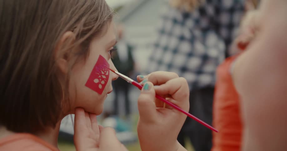 A close-up over-the-shoulder shot of a young girl having a floral design painted on her cheek using a stencil at an outdoor event