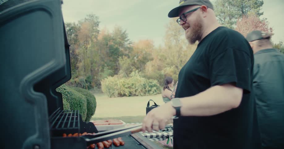 A man with a beard and glasses smiles while grilling sausages at a backyard barbecue on a sunny day