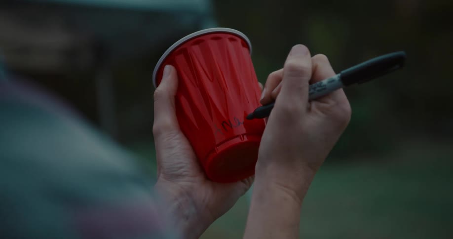 A person's hands write a name on a red plastic party cup with a black marker during an outdoor gathering