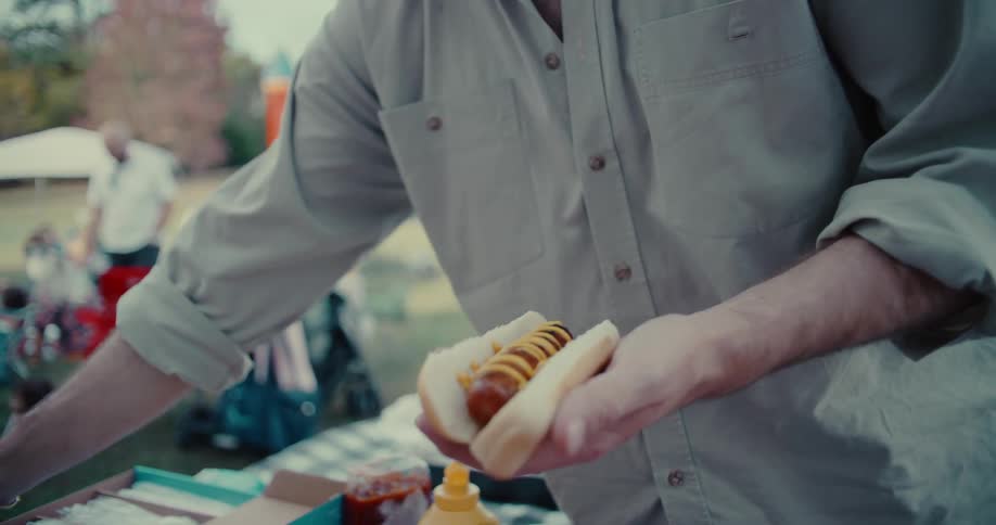 A man prepares a hot dog in slow motion with ketchup and mustard at a lively outdoor party