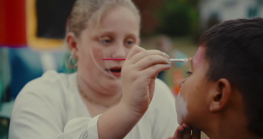 A focused young girl carefully applies colorful face paint to a boy's forehead at an outdoor event
