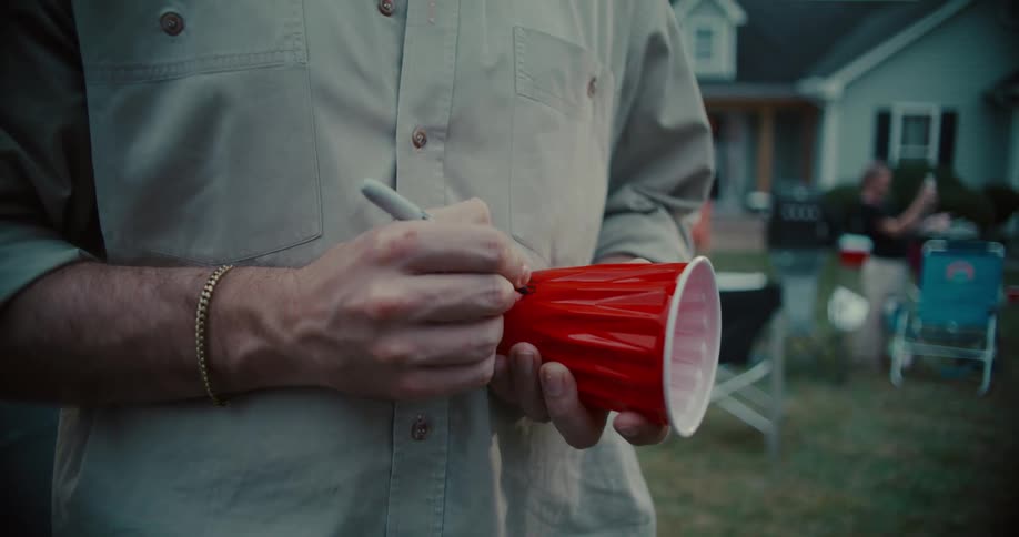 A young man writes his name on a red plastic cup with a marker at an outdoor party