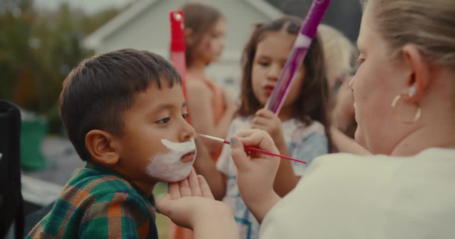 A woman carefully paints a young boy's face with white makeup at a children's party outdoors