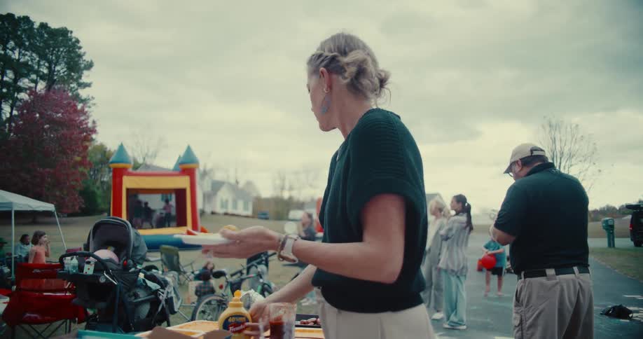A woman carefully adds mustard to her plate at a lively neighborhood block party on a cloudy day