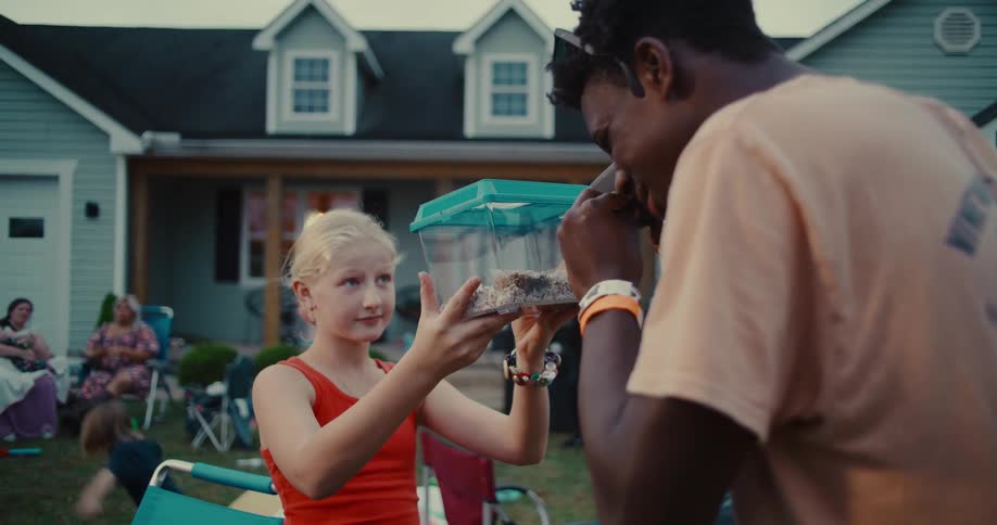 A young girl holds up a bug terrarium for a young man to inspect as they hang out in the front yard during a neighborhood gathering