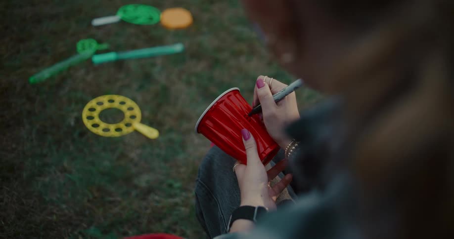 An over-the-shoulder view shows a person writing their name on a red plastic cup at a backyard party