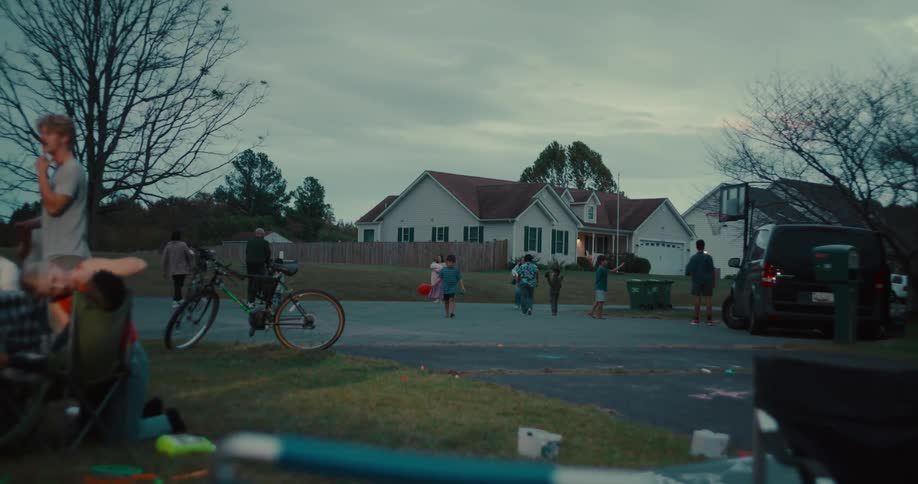 A diverse group of neighbors and children gather to play basketball and socialize in a suburban cul-de-sac at dusk