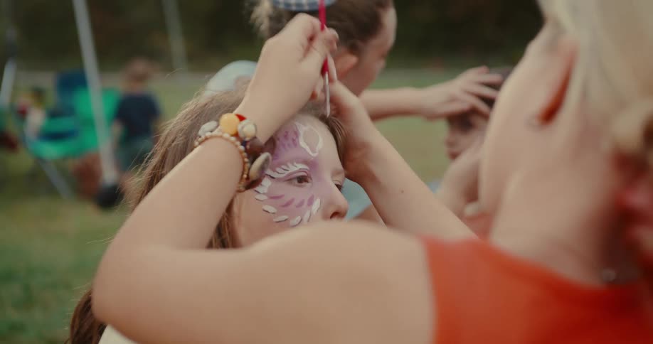 A patient young girl gets a beautiful purple and white butterfly design painted on her face by an artist at an outdoor party