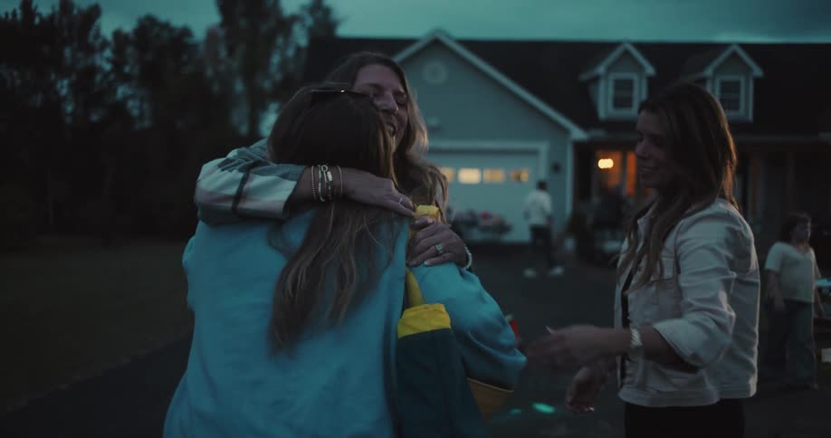 Two women welcome a friend with a warm hug as she arrives at a block party in their neighborhood at dusk