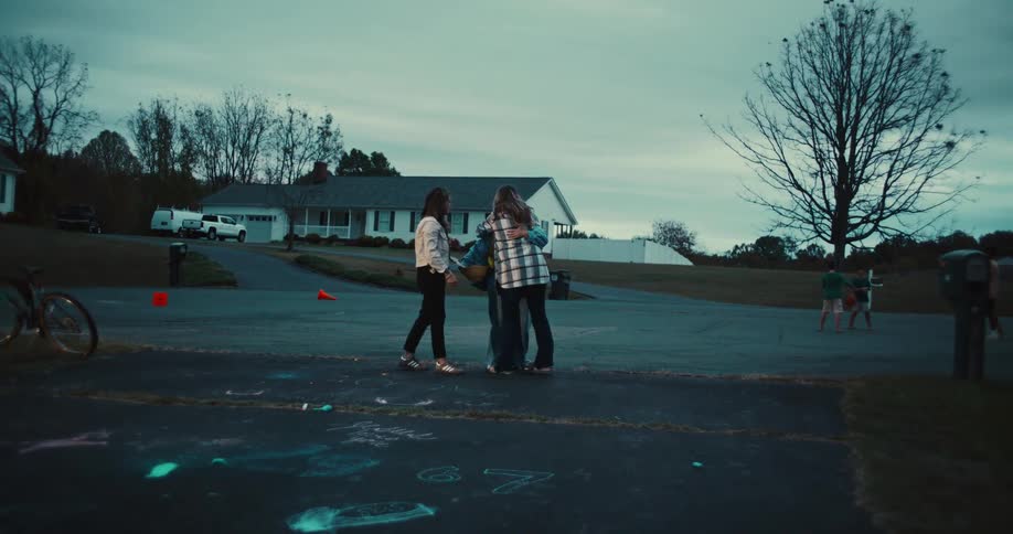 Three women hug, laugh, and walk down a driveway together in a suburban neighborhood at dusk