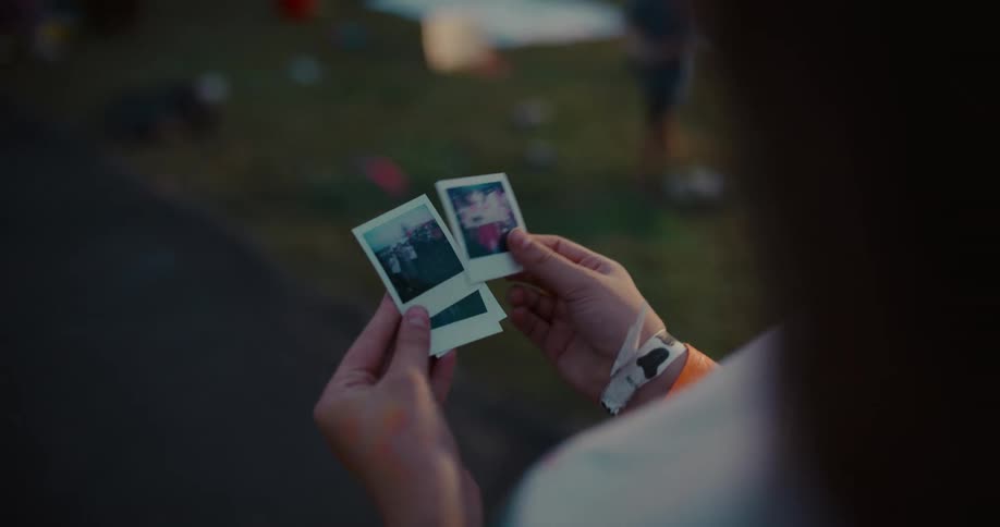 A person with festival wristbands fondly looks through instant photographs of friends and memories in a nostalgic slow-motion shot