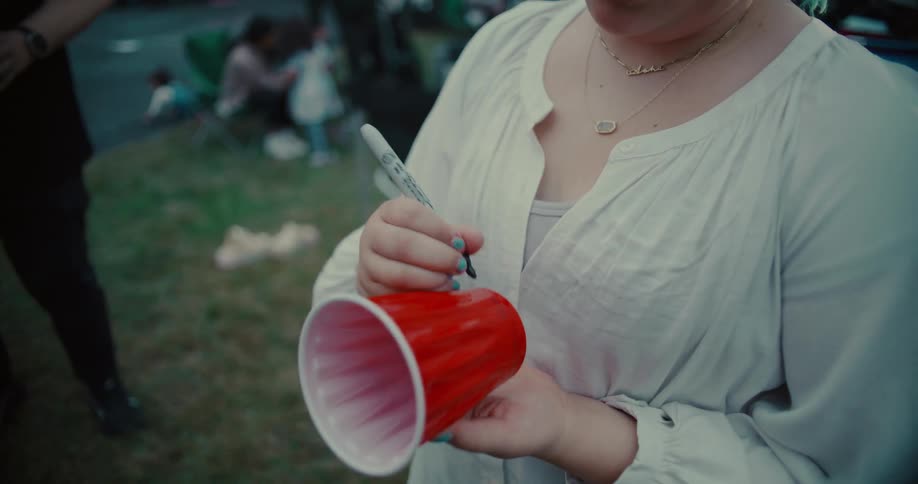 A young woman writes her name on a red plastic cup with a permanent marker at an outdoor party in slow motion