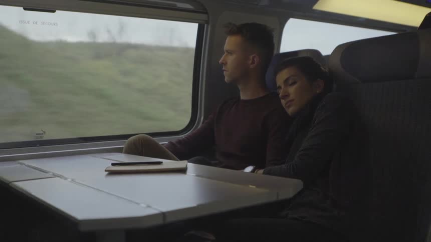 A man looks out a train window as a woman rests while traveling