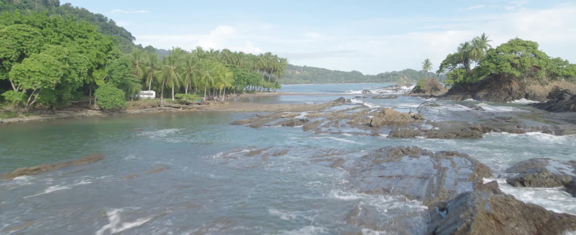 Drone slowly flies over a rocky tropical coastline toward a secluded beach lined with lush palm trees