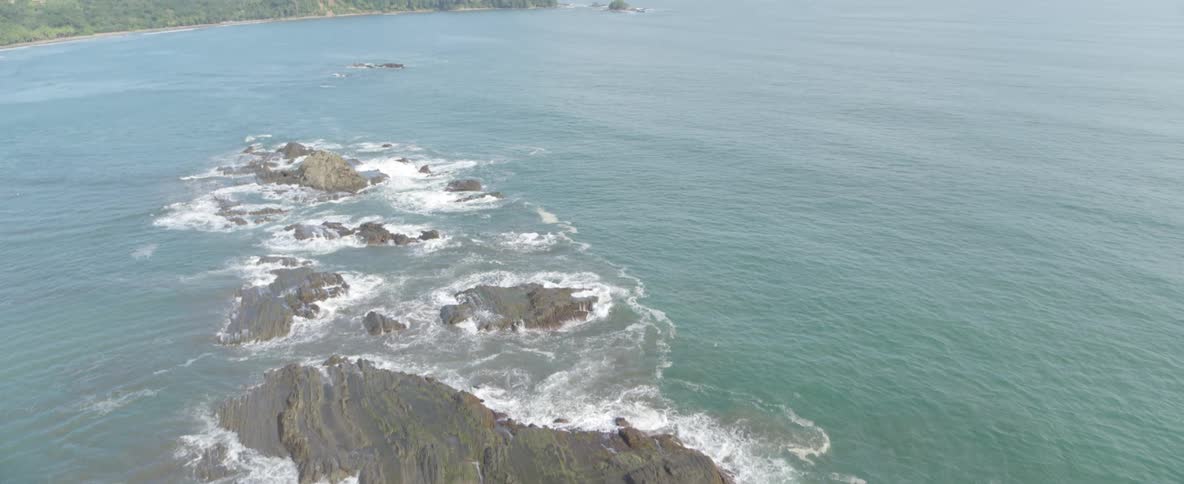 A slow aerial reveal of a tropical rocky coast with gentle waves breaking on the formations and a distant jungle-covered shoreline