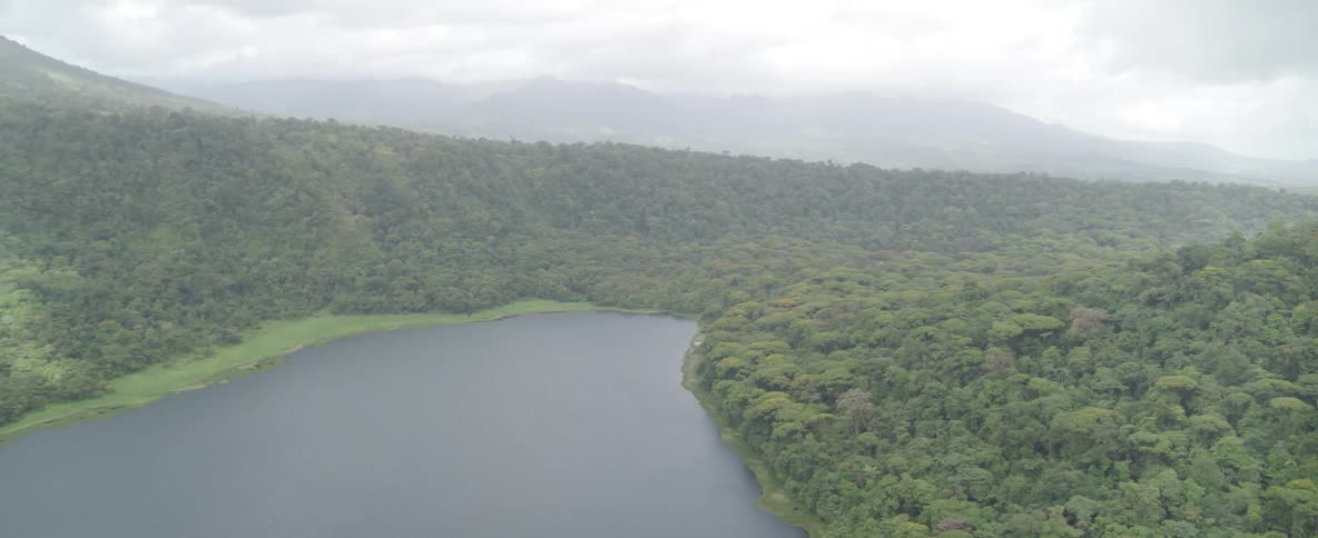 A slow, pulling-back aerial shot reveals a tranquil lake surrounded by a lush green rainforest and distant misty mountains