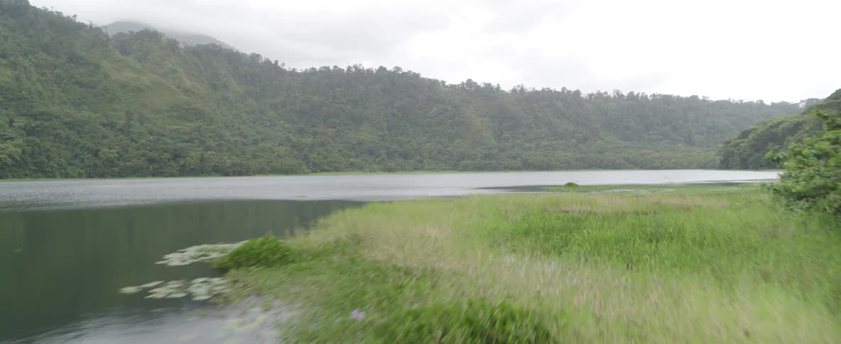 A serene aerial view flying low over the still waters of a remote lake surrounded by lush green mountains on an overcast day