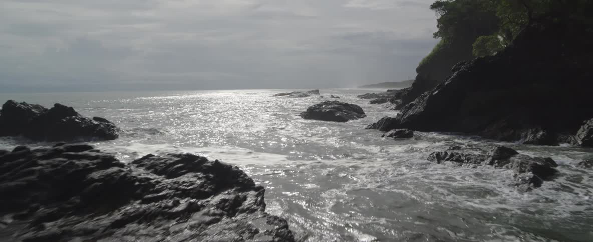 Aerial drone shot pulls back low over a rocky coastline as waves crash on a bright, overcast day