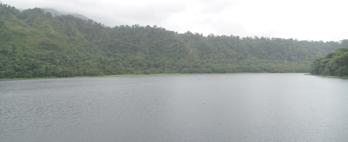 Light rain falls on a serene lake nestled at the base of a dense, forested mountain