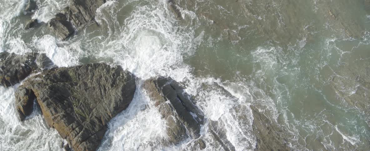 An aerial top-down view shows powerful ocean waves crashing against a rocky shoreline in slow motion