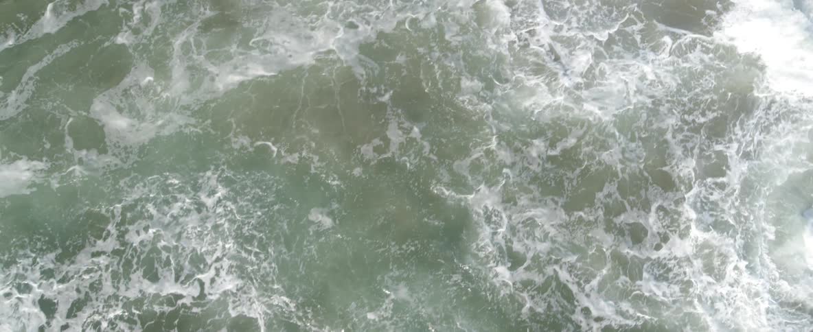 Mesmerizing top-down aerial footage shows foamy ocean waves swirling around and crashing against large coastal rocks in slow motion