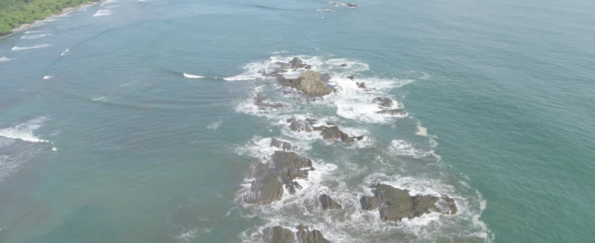 Aerial view slowly pulls back to reveal a rocky peninsula with palm trees on a tropical coastline