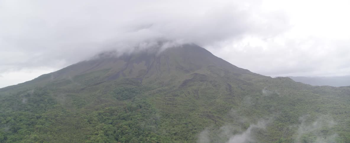 A static aerial shot captures the misty green slopes of a large volcano, its peak hidden by a thick blanket of clouds