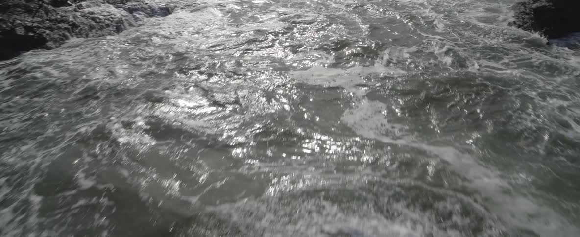 The camera pulls back over shimmering, choppy ocean water to reveal a rugged, rocky coastline