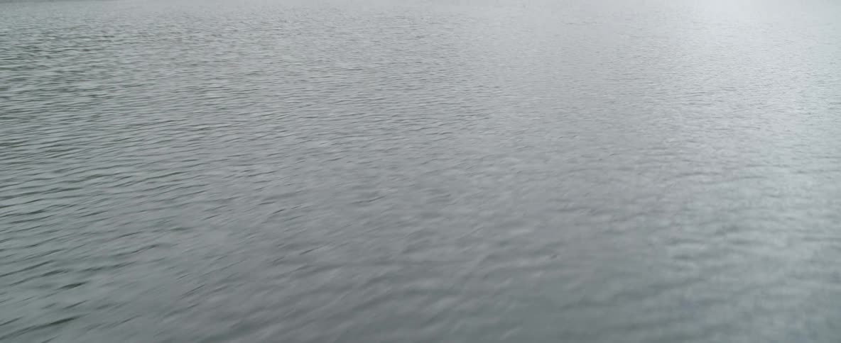 A slow tilt up from the water's surface reveals a calm lake surrounded by lush green mountains on an overcast day