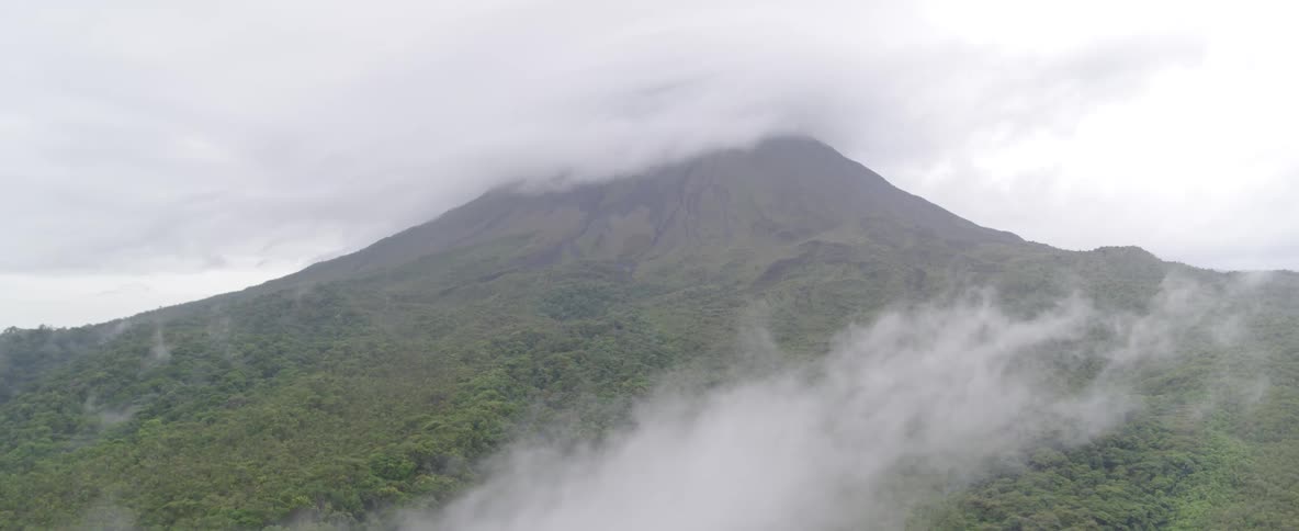 A majestic, cloud-covered volcano stands tall over a lush green forest on a misty day