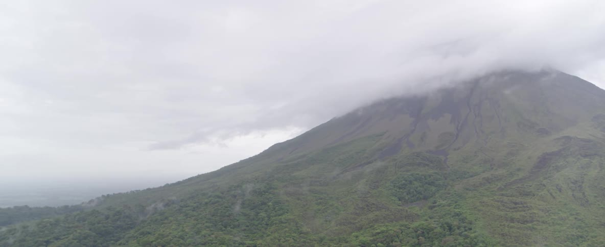 An aerial shot slowly pans across a majestic volcano with its peak shrouded in clouds, revealing a vast jungle landscape below