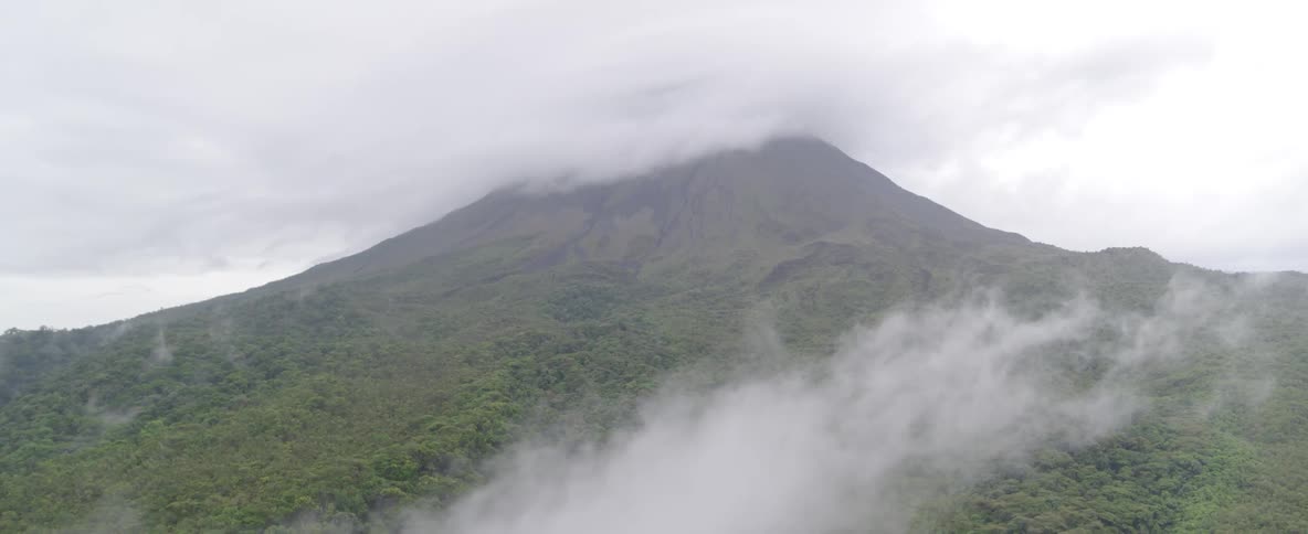 A static aerial view shows a majestic green volcano with its peak shrouded in a thick layer of clouds on a misty day