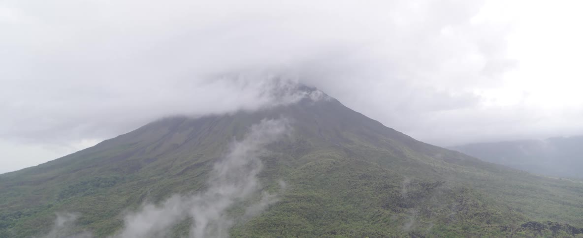 A static aerial view shows a massive, green volcano with its peak completely covered by thick, rolling clouds